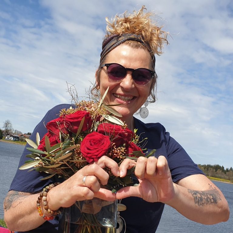 Lisa, Licht-im-ich.de Frau mit Lockenfrisur und Sonnenbrille, hält einen Blumenstrauß und bildet ein Herz mit den Händen.